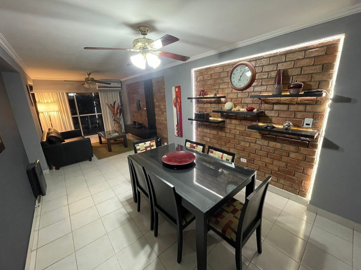 A dining area features a sleek black table surrounded by colorful chairs. A brick wall is decorated with floating shelves displaying decorative items. Ceiling lights and a standing lamp create a cozy ambiance, while the adjacent living room is visible through an open space.