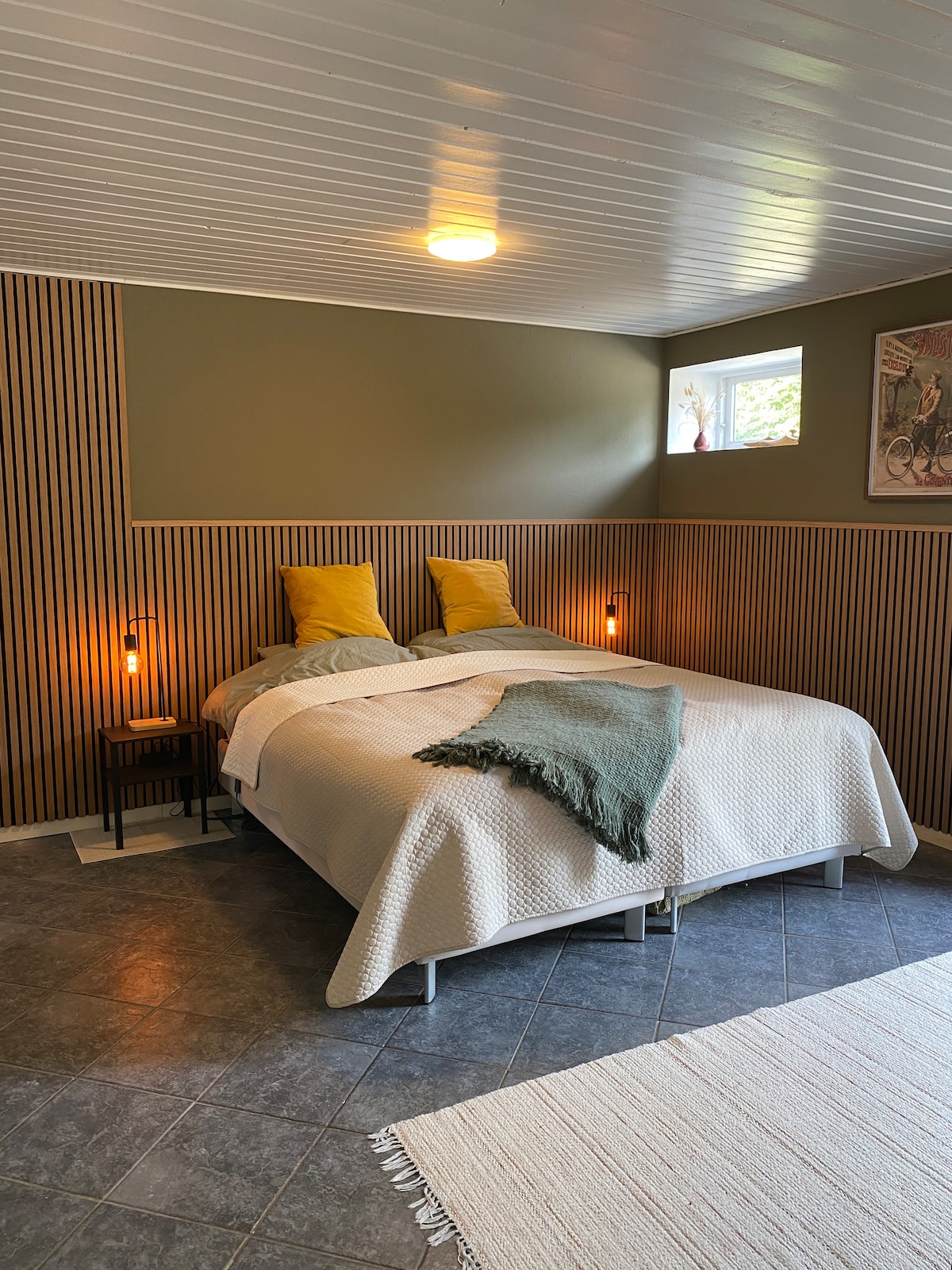 A sleeping area features a spacious bed with a textured blanket draped across the foot. Two yellow pillows are positioned against a green accent wall, while natural light filters through a small window. Warm lighting is provided by wall sconces on either side of the bed.