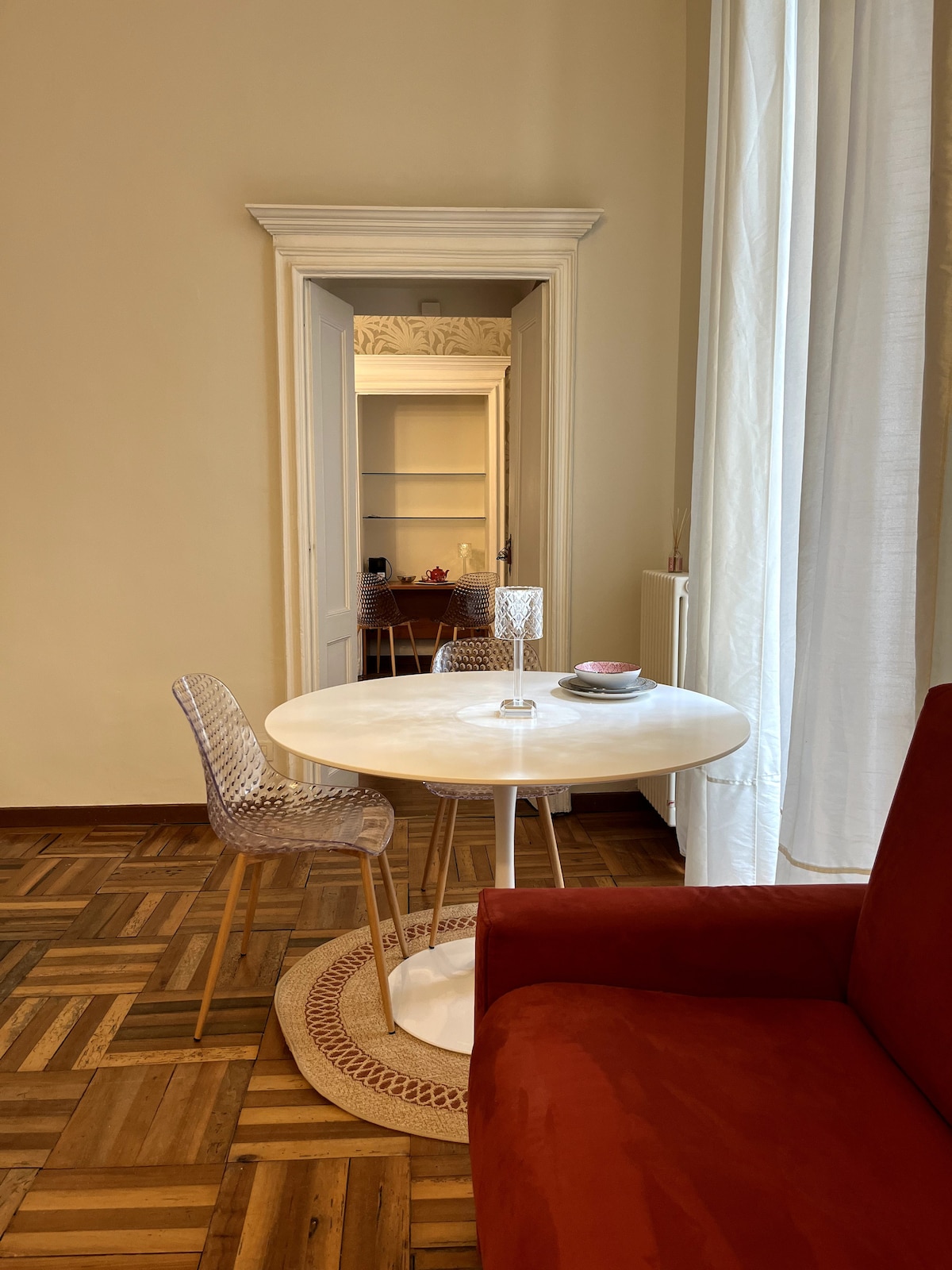 A round white dining table is set with plates and a decorative candle holder, accompanied by transparent chairs. A red couch is positioned nearby, and a doorway in the background leads to a room with a softly lit ambience, complemented by white drapes allowing natural light to enter.