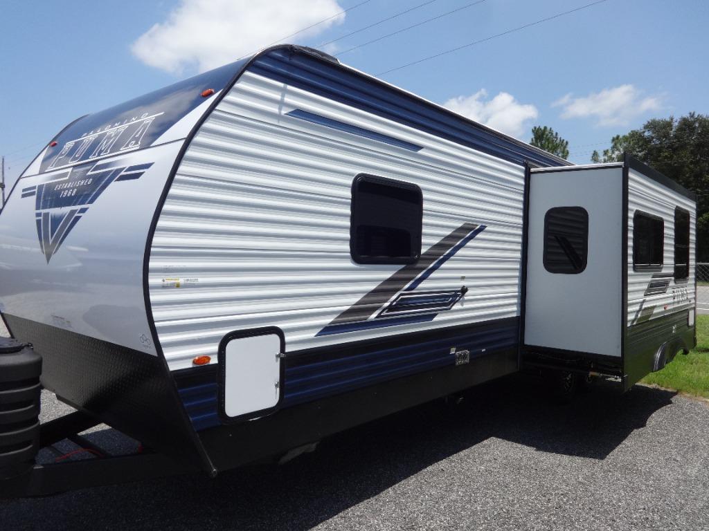 The exterior of a modern RV is displayed, featuring a streamlined design with a combination of blue and silver accents. A slide-out section on one side adds extra space, while large windows allow for natural light. The RV rests on a gravel surface, surrounded by greenery.