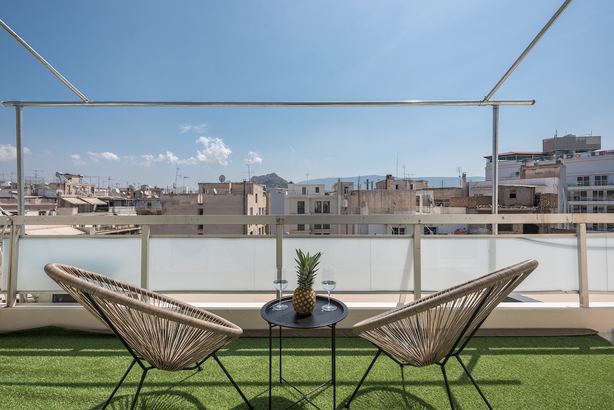A private balcony is equipped with two woven chairs facing a small table, featuring a pineapple and two glasses. The space is draped in artificial grass and overlooks a cityscape under a clear blue sky.