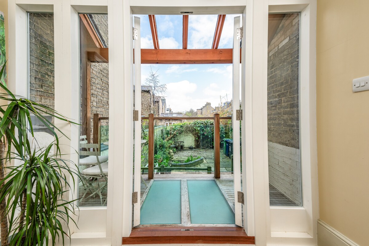 Double glass doors open to a serene outdoor space, framed by wooden beams. Lush greenery can be seen in the garden, complemented by a wooden deck and subtle seating arrangement. Natural light enhances the inviting atmosphere.
