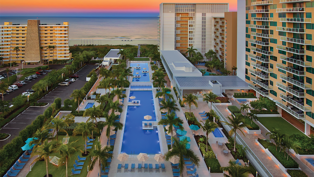 The expansive outdoor pool area features a long, inviting pool surrounded by palm trees and lounge chairs. The ocean can be seen in the background, with sunset hues illuminating the sky. Well-maintained pathways lead to various amenities spread throughout the landscaped grounds.