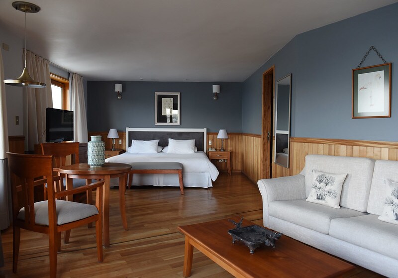 A spacious suite features a Super King bed and an individual bed, complemented by a comfortable sofa and wooden dining set. Wooden accents adorn the room, and natural light enters through the large window. A flat-screen television and bedside lamps are included.