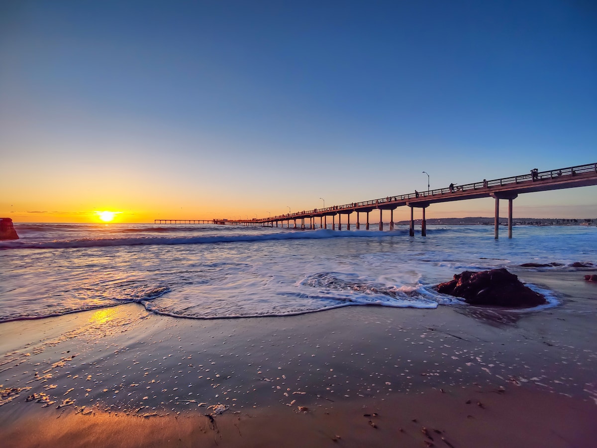The image captures a serene beach scene at sunset, where waves gently lap at the shore. A distant pier extends into the ocean, with soft hues of orange and blue reflecting off the water. The shoreline is highlighted by wet sand and scattered pebbles.