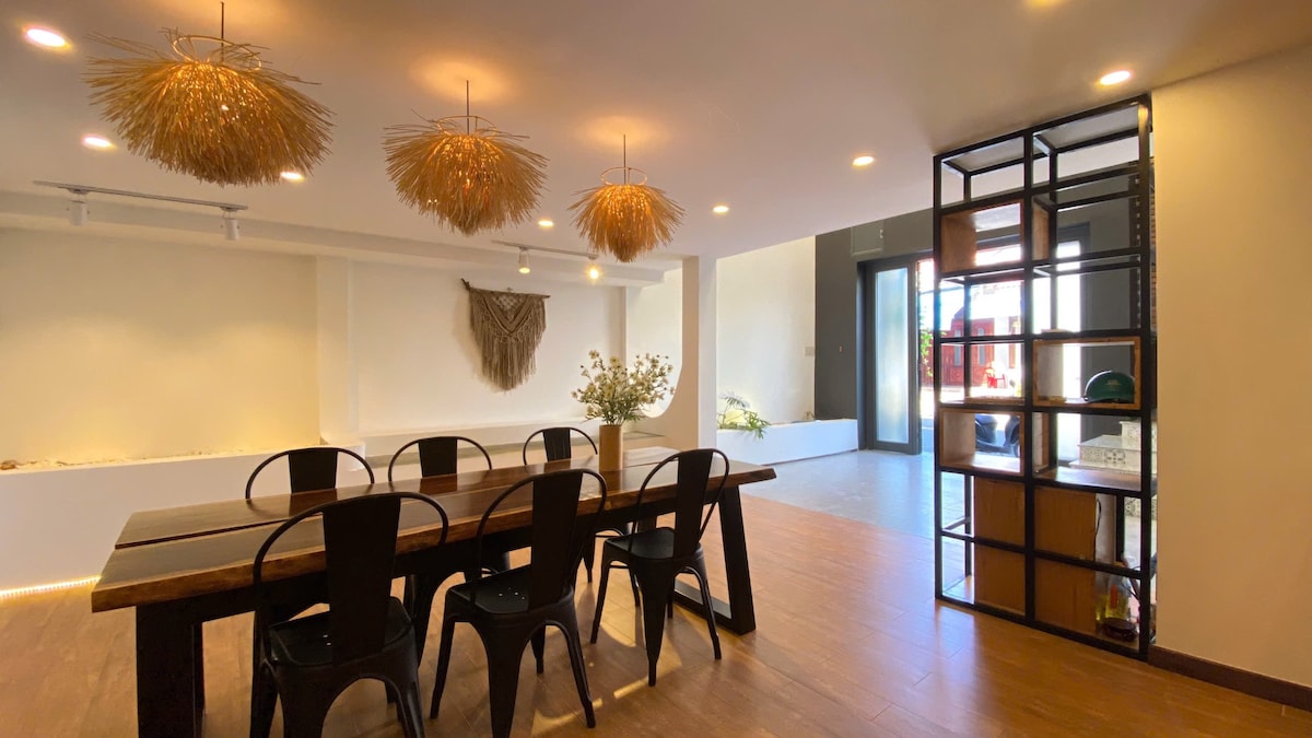A spacious dining area is highlighted by a large wooden table surrounded by black chairs. Unique straw light fixtures hang from the ceiling, adding character. A decorative wall piece is present, and bright natural light flows through a nearby door, creating an open and welcoming atmosphere.