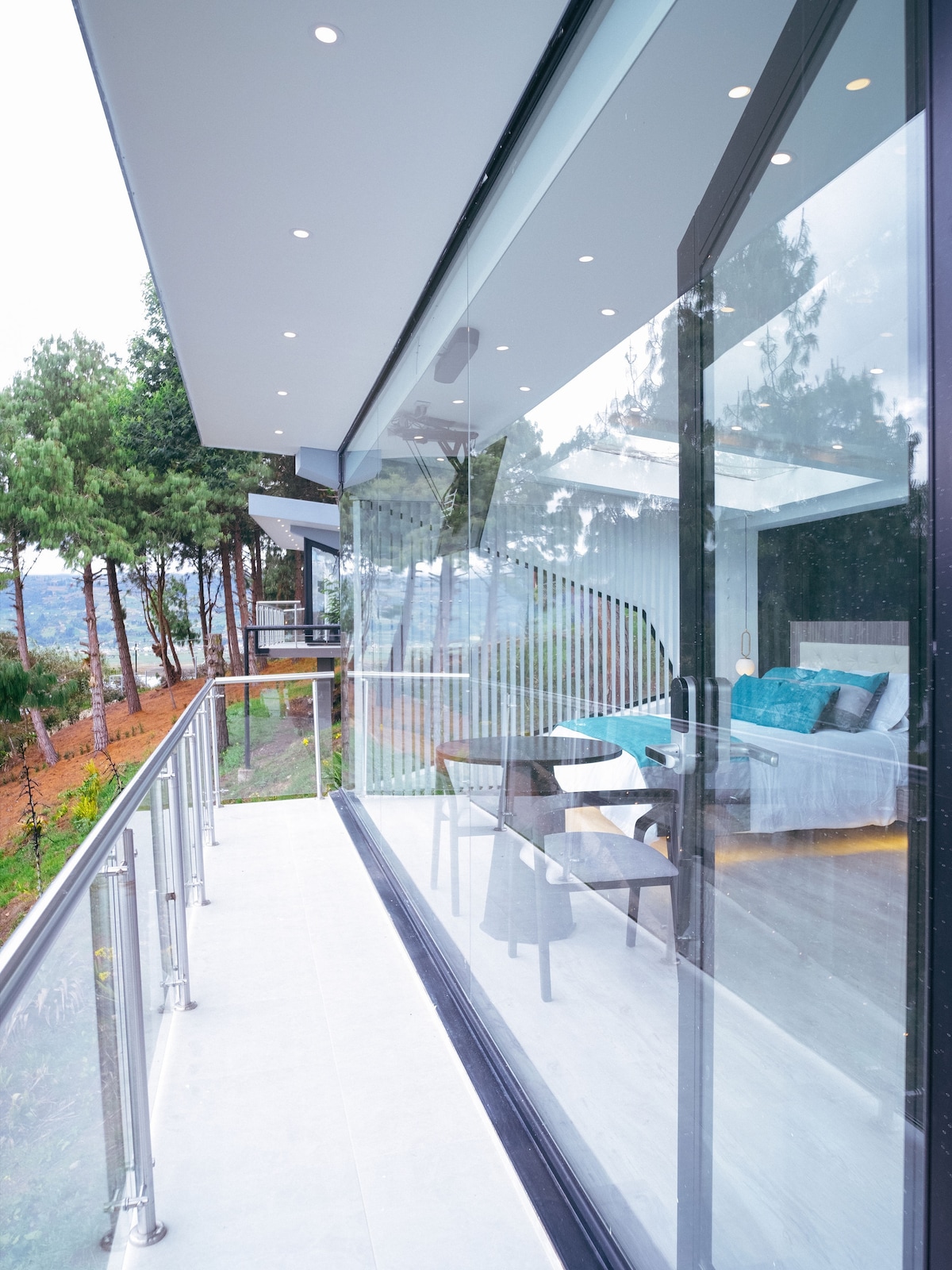 A modern space features floor-to-ceiling glass walls, providing extensive views of the surrounding greenery. A comfortable seating area with a small table is visible. Light-colored walls and decorative elements contribute to a serene ambiance.