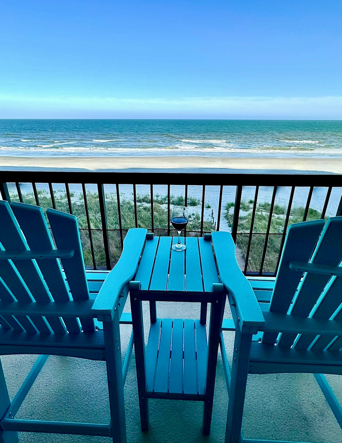 A private balcony features two bright blue chairs flanking a small table. A glass rests on the table, overlooking an expansive view of the ocean and sandy beach, with gentle waves lapping at the shore under a soft blue sky.