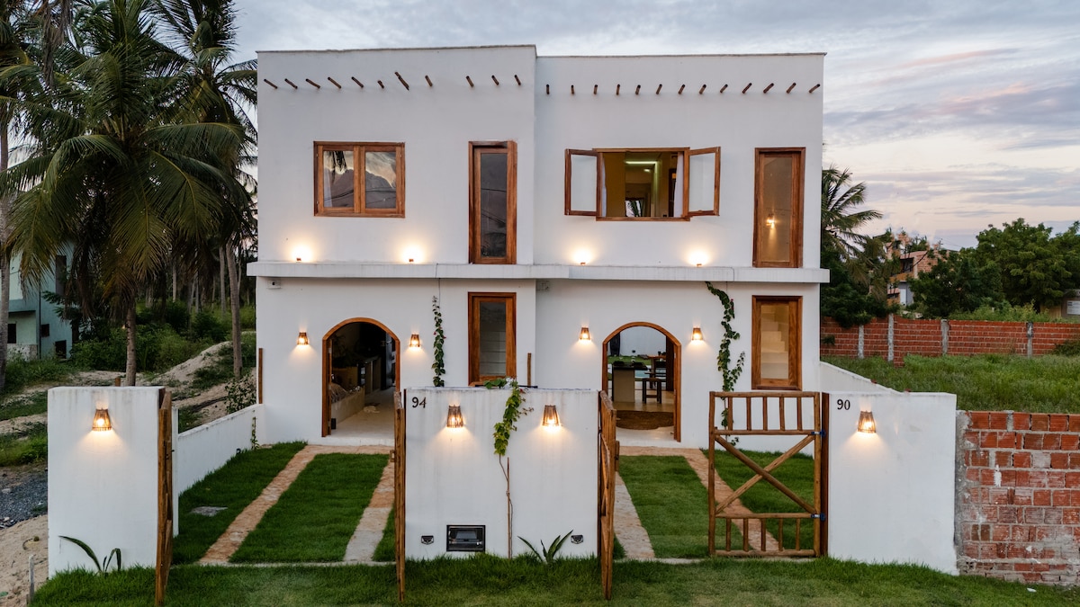 The exterior of a two-story house is presented, showcasing a minimalist design with white walls and wooden accents. Symmetrical windows and illuminated arches provide a welcoming look. Lush green lawns lead to a rustic wooden gate, enhancing the inviting atmosphere of the property.