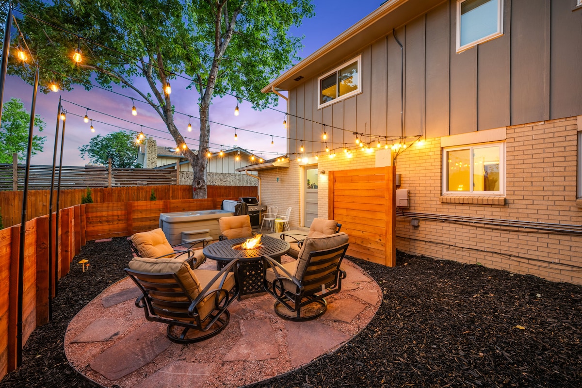 A fenced outdoor space is highlighted by a circular stone patio with comfortable seating around a fire pit. Warm string lights create a cozy atmosphere above the area, featuring a hot tub and grill, surrounded by freshly mulched landscaping.