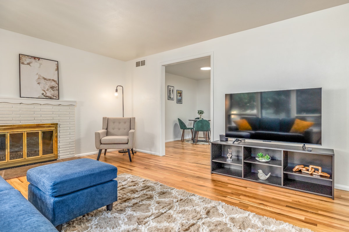 The living area features a comfortable seating arrangement with a blue sectional sofa and a modern armchair. A flat-screen television is positioned on a black shelving unit, with a stylish light fixture beside the chair. Warm wooden floors add to the cozy ambiance.