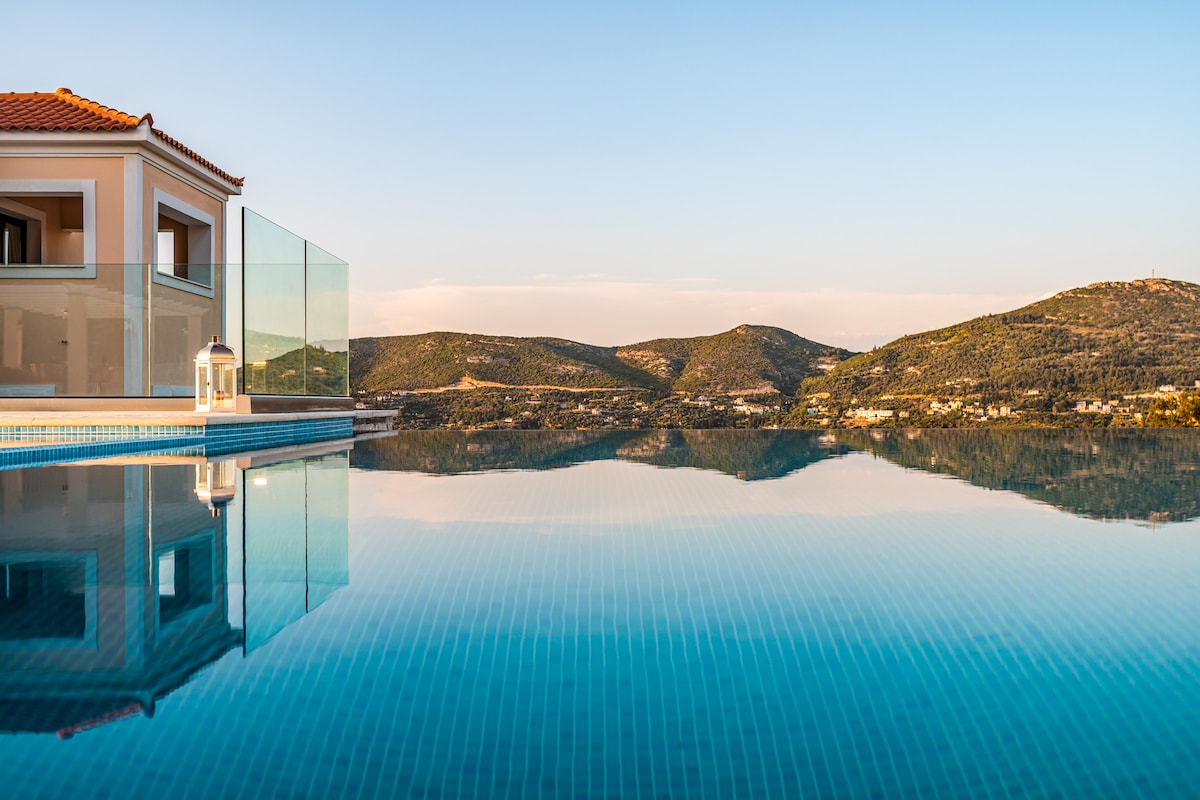 An infinity pool reflects the surrounding mountains and sky, creating a seamless horizon. The tranquil water complements the modern architecture of the villa, with a glass railing enhancing the view. Soft colors of the landscape are captured in the calm surface of the pool.