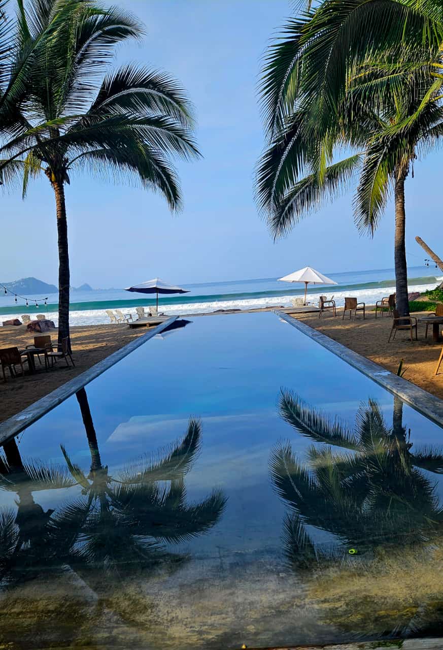 An infinity pool is situated in the foreground, reflecting tall palm trees and the expansive blue sky. Beach chairs and umbrellas are visible along the sandy shore, leading to the gentle waves of the ocean in the background.
