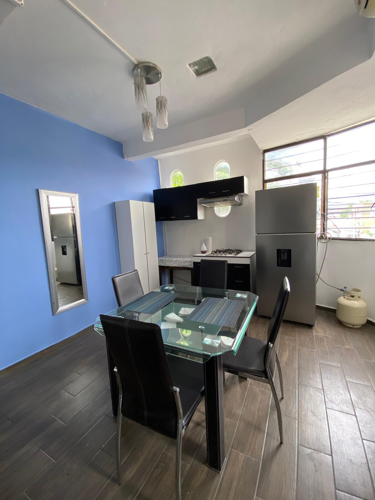 An airy kitchen area features a glass dining table surrounded by four black chairs. Modern appliances are present, including a refrigerator and a sleek white cabinet. Large windows allow natural light to illuminate the space, highlighting the blue accent wall and bringing warmth to the atmosphere.