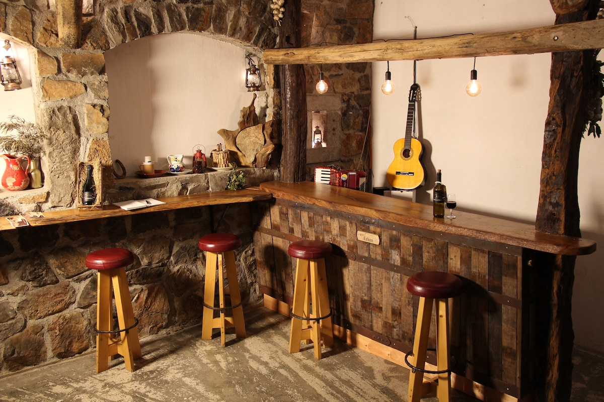 A rustic bar area features a wooden counter supported by sturdy wooden beams. Four stools with red cushioned seats are arranged along the counter. Ambient lighting is provided by hanging bulbs, and decorative items are displayed on the shelves along the stone wall.