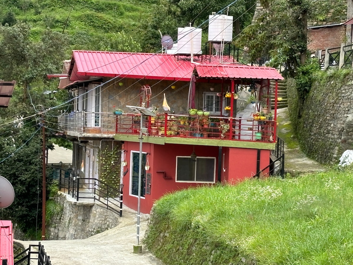 A two-story building with a red roof and exterior features a blend of stone and bright red walls. The upper level includes a spacious terrace adorned with potted plants. Nearby pathways are lined with greenery, creating a serene mountain ambiance.