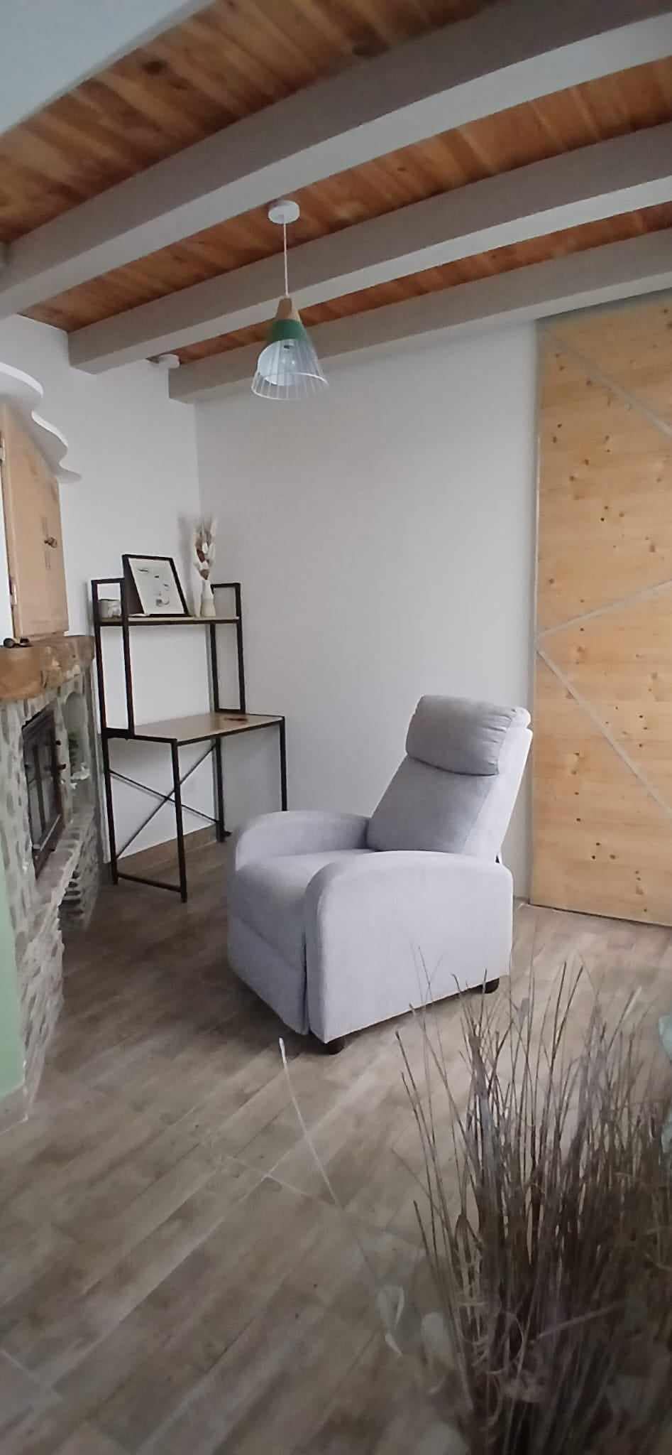 A light-filled corner features a soft, upholstered armchair beside a minimalist black shelving unit. The stone fireplace is adorned with decorative frames, while natural wood accents from the ceiling and floor complement the neutral color palette of the room.