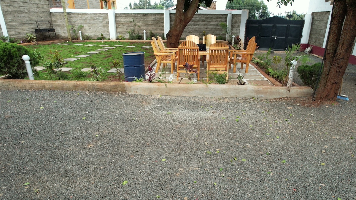 An outdoor seating area is visible, featuring a wooden dining table surrounded by six matching chairs. A well-maintained garden space is seen in the background, with neatly arranged pathways and greenery. The gravel surface adds a rustic charm to the setting.
