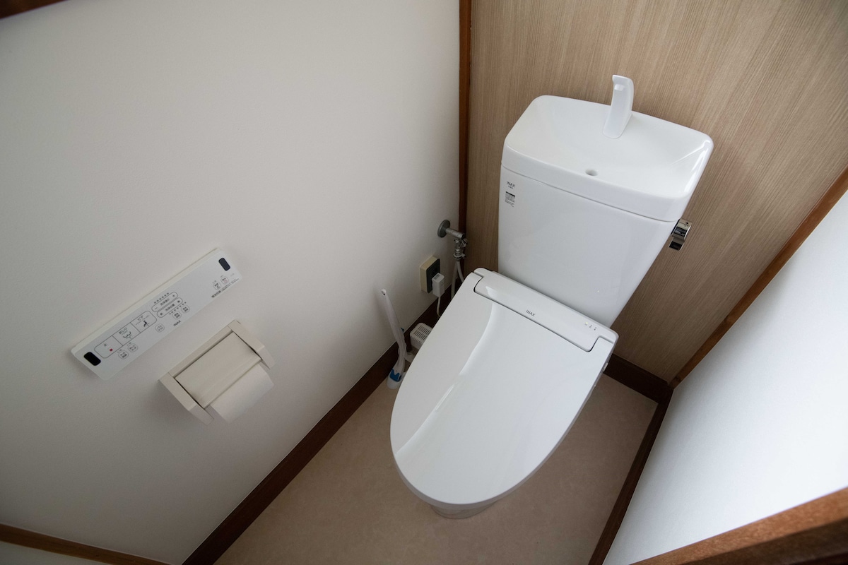 The image shows a modern bathroom featuring a clean, white toilet with a sleek design. A control panel is mounted on the wall nearby, and a toilet paper holder is discretely installed. The surrounding walls are finished with light-colored materials, providing a simple and functional aesthetic.