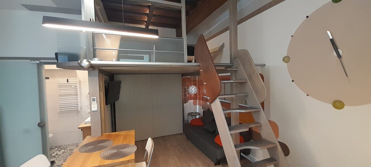 The interior of the studio features a wooden staircase leading to a sleeping loft. A dining table with four chairs is situated beside a gray sofa, accented with colorful cushions. Decorative elements include a round wall clock and vibrant wall art.