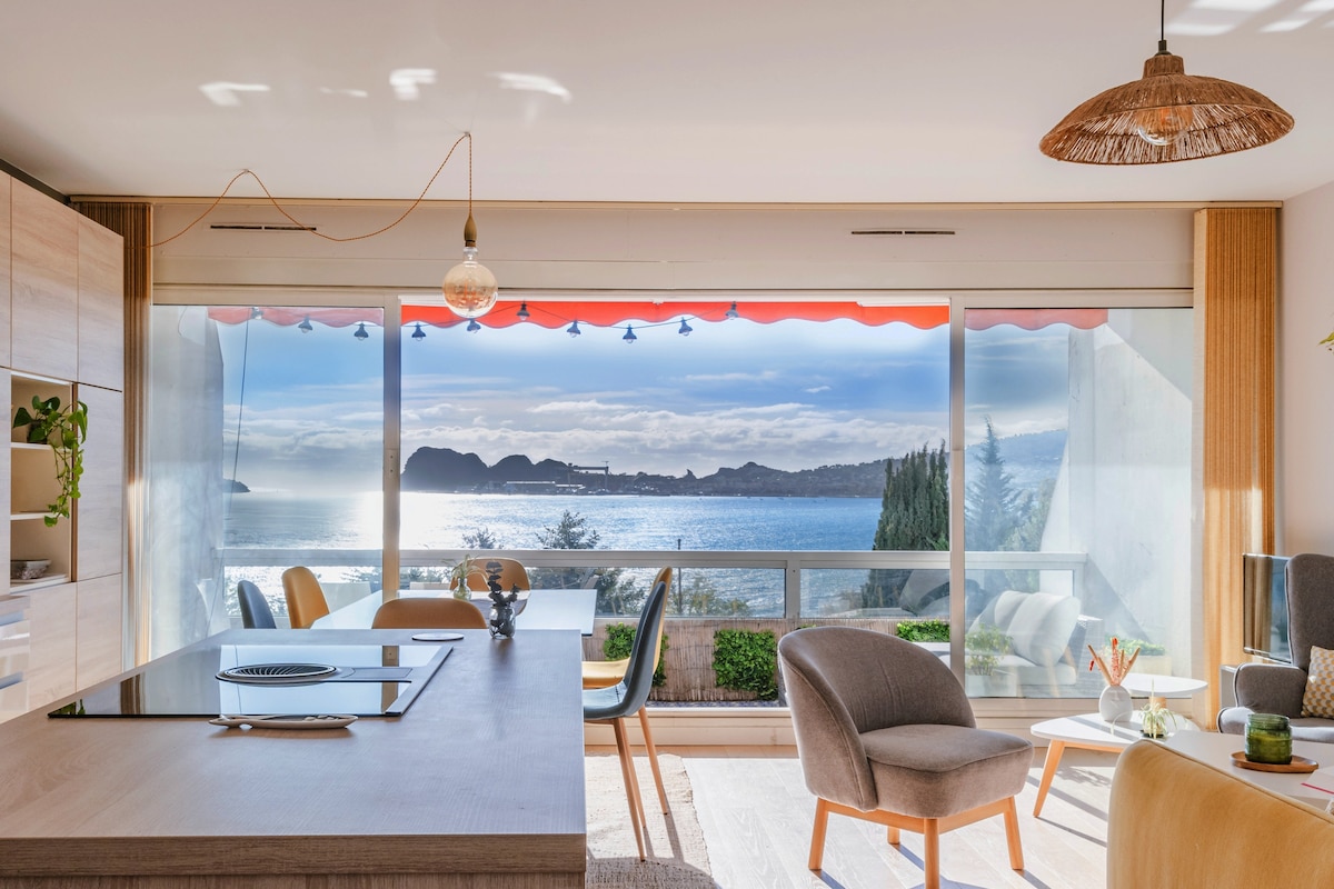 An open living space features a modern kitchen and dining area with a large window offering expansive sea views. Natural light floods the room, highlighting light wood accents and greenery. A comfortable seating area is visible, creating a harmonious atmosphere with the outdoor scenery.