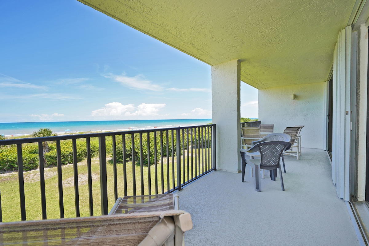 A private balcony is shown overlooking lush greenery and the Atlantic Ocean. Four plastic chairs are positioned for relaxation, while the shoreline can be seen in the distance under a bright blue sky. The space is illuminated by natural light.