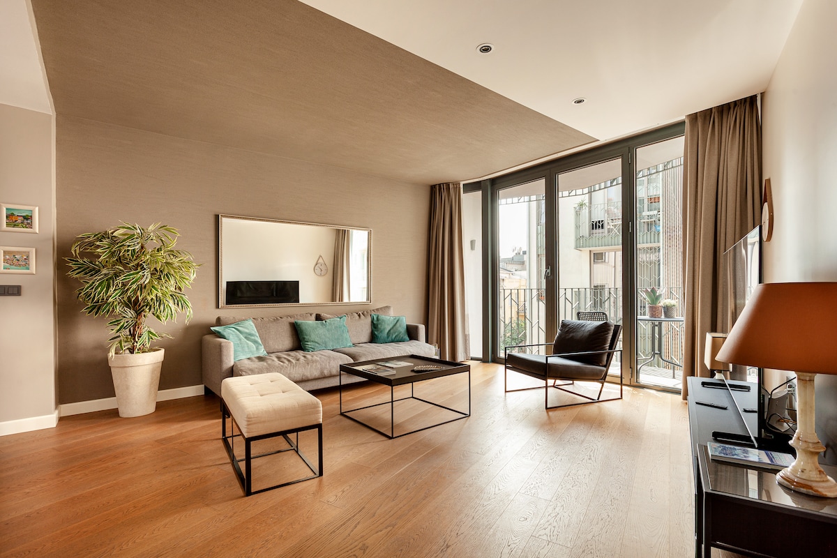 The living area features a modern sofa paired with a stylish armchair, both arranged around a minimalist coffee table. Natural light floods the room through large windows, with a view of the balcony. A potted plant adds a touch of greenery to the space.