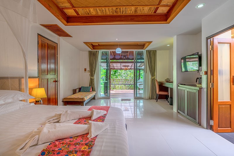 An air-conditioned room features a spacious bed adorned with decorative linens. Natural light fills the space through large glass doors that open to a terrace. A seating area with a bench and a flat-screen TV are also present, contributing to a relaxed atmosphere.