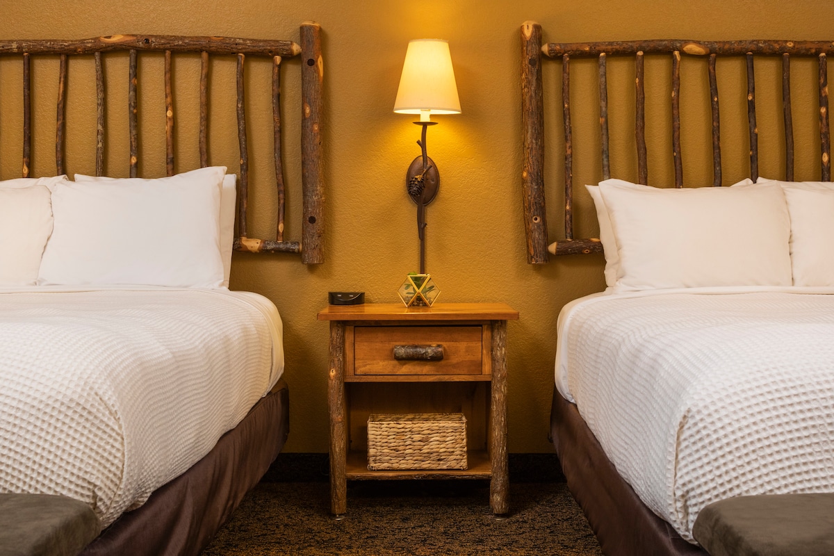 Two spacious beds are framed by rustic wooden headboards, crafted from natural materials. A simple wooden nightstand with a woven basket sits between the beds, topped with a yellow decorative piece and a lamp that casts a soft glow in the warm-hued room.