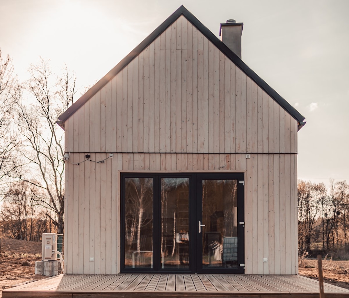 The rear view of the wooden house presents large glass doors that open to a spacious terrace. The natural wood exterior contrasts with the clear sky, while surrounding trees enhance the serene setting. A chimney rises above the gabled roof.