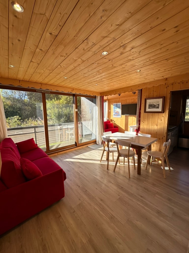 A sunlit living area features wooden ceilings and large windows providing natural light. A red sofa complements a simple white dining table surrounded by four chairs. The balcony offers a view of the outdoors, enhancing the overall spaciousness of the room.