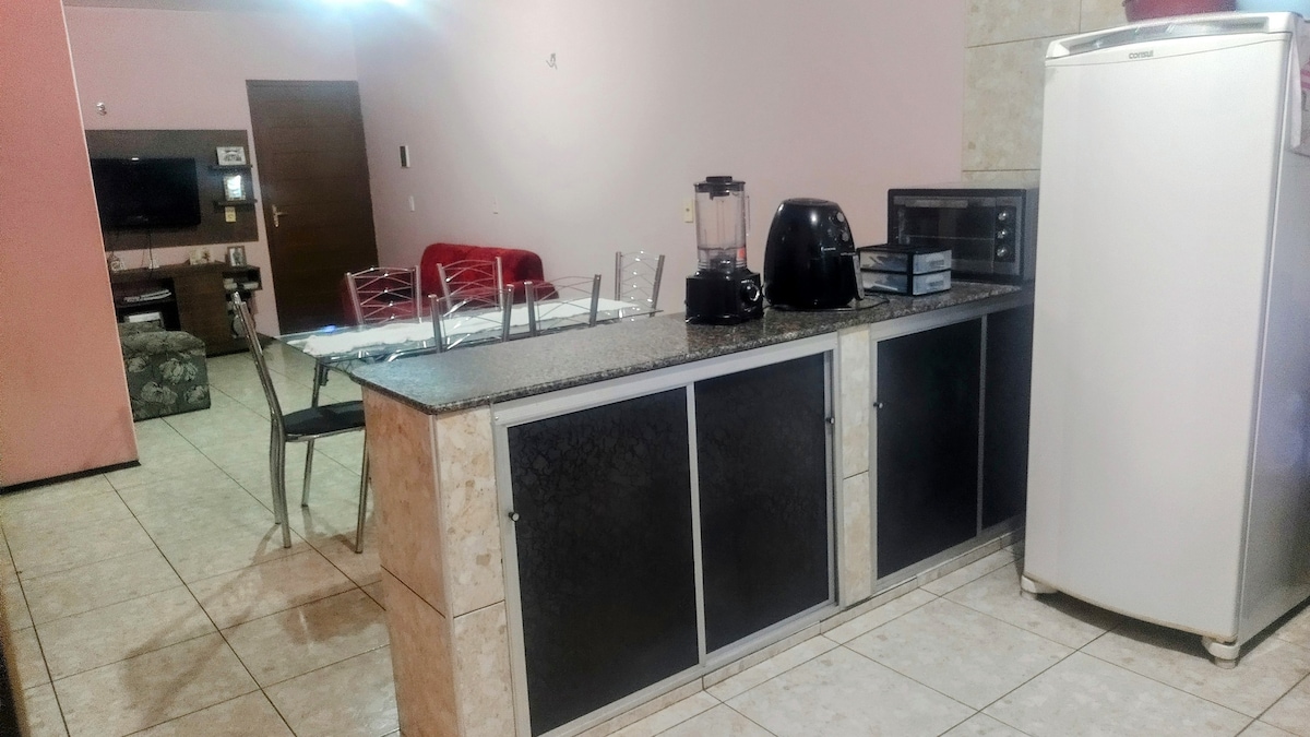 The kitchen area features a granite countertop with a black and silver blender, a coffee maker, and a microwave. A white refrigerator stands nearby. Four metal chairs are positioned around a dining table, with light tile flooring completing the space.