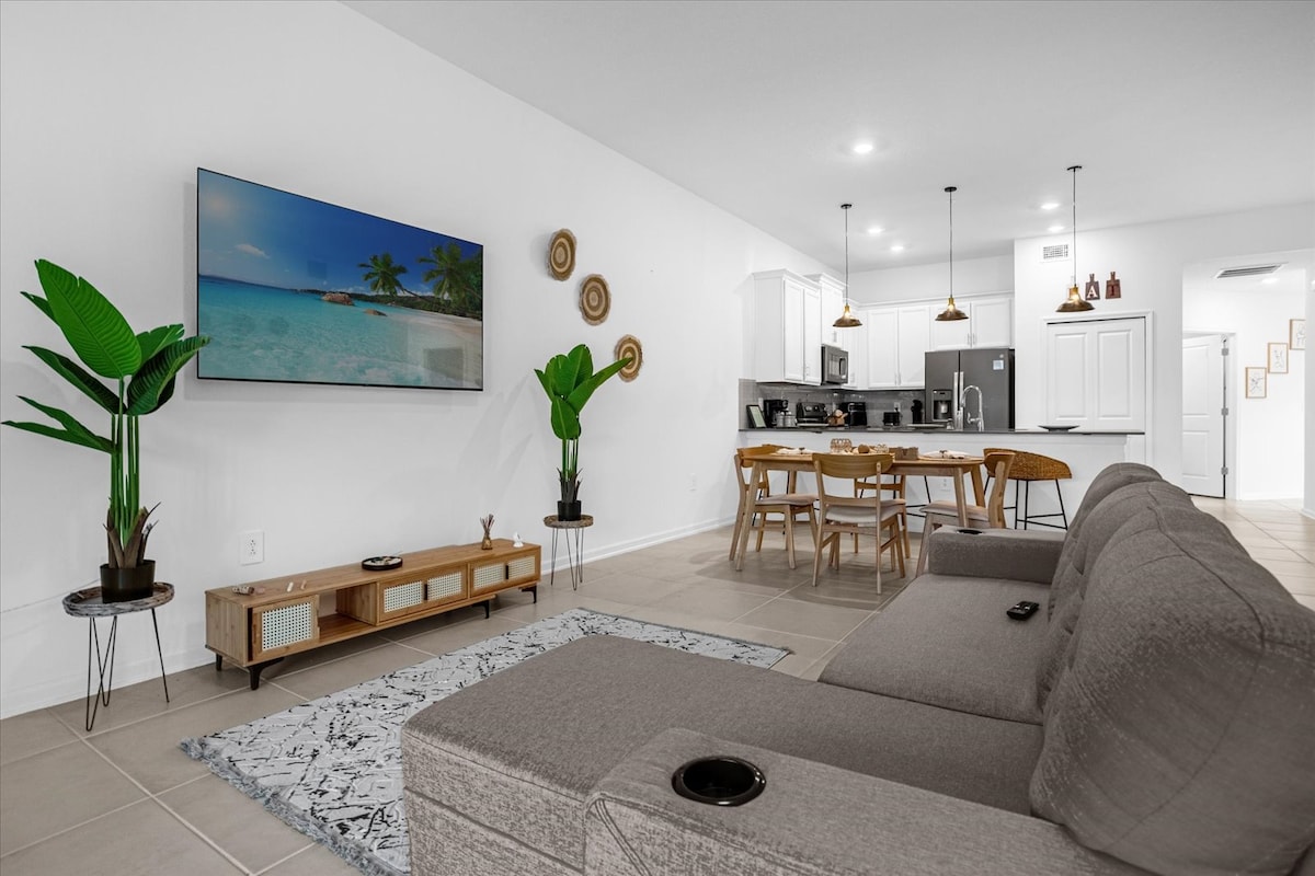 A spacious living area features a comfortable sectional sofa with built-in cup holders. A large television is mounted on the wall, providing entertainment. A dining table with stylish chairs is positioned nearby, and potted plants add a touch of greenery to the neutral decor.