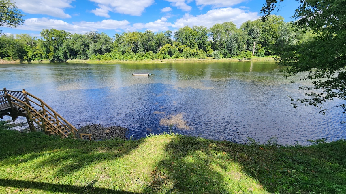 A serene view of the Potomac River features calm waters reflecting the blue sky and scattered clouds. Lush green trees line the riverbanks, providing a natural backdrop. A wooden deck with stairs leads from the grassy area to the water's edge, inviting relaxation and outdoor activities.