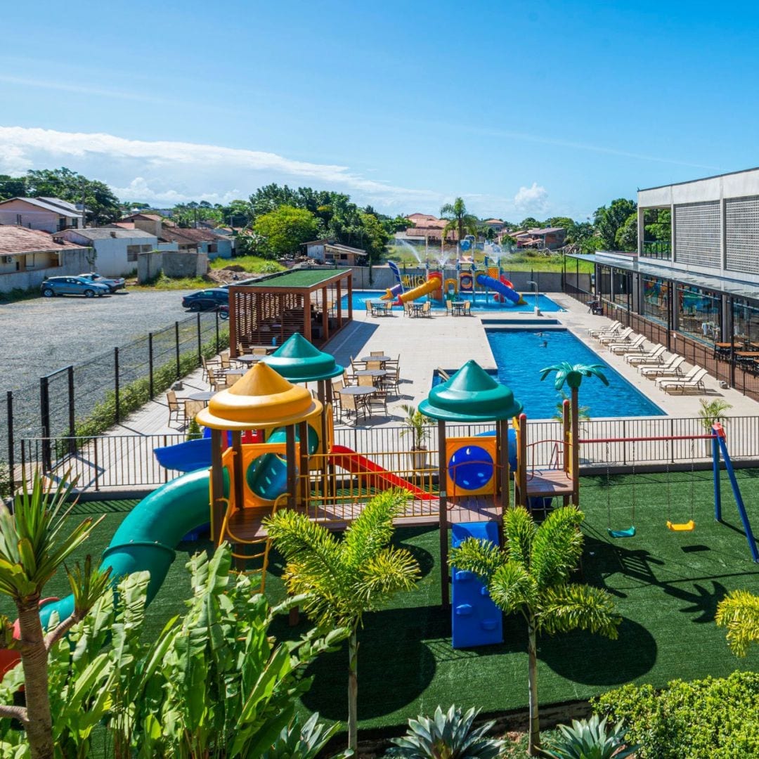 An outdoor area is presented featuring a swimming pool surrounded by sun loungers. Colorful playground structures are visible, including slides and climbing frames. Lush green landscaping surrounds the space, creating an inviting environment for families and children.