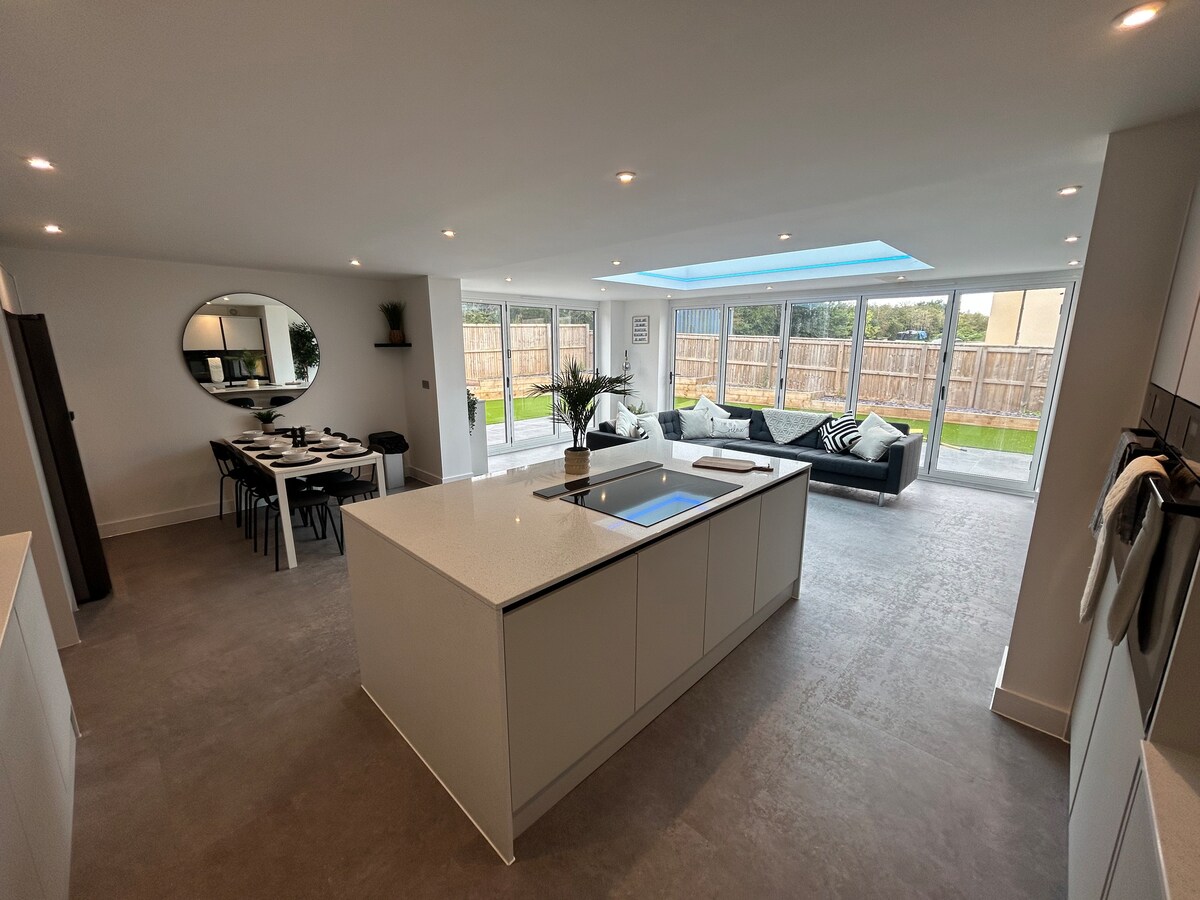 A modern kitchen area features an island with a built-in sink and seating, surrounded by sleek cabinetry. Natural light fills the space, highlighted by a skylight above. A dining table is visible in proximity, with a comfortable seating area composed of a sectional sofa and decorative plants.