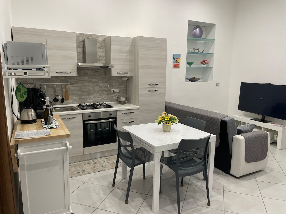 A bright kitchen and living area features light-colored cabinetry and a gas stove. A dining table with four chairs sits beside a gray sofa. A television is positioned in the corner, while shelves display decorative items. White tiled flooring enhances the spacious feel.