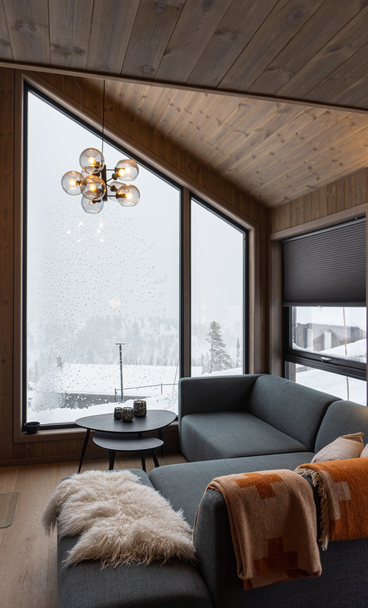 A modern living area features large, angled windows that frame a snowy landscape outside. A soft, textured blanket drapes over a sectional sofa, complemented by a minimalist coffee table holding decorative elements. Warm lighting from a uniquely designed fixture enhances the cozy ambiance.