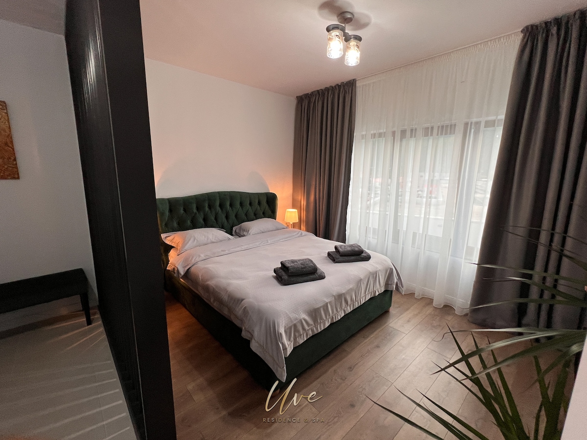 A spacious sleeping area features a plush green bed frame with white bedding and matching towels. Soft lighting is provided by a bedside lamp, while large windows are adorned with sheer curtains and darker drapes, allowing natural light to soften the room.