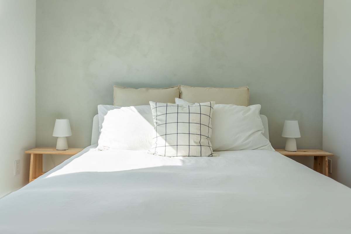 A spacious bed is presented with soft, neutral linens. Two bedside lamps are positioned on wooden nightstands, providing a balanced aesthetic. The light, airy decor is complemented by a textured wall, creating a soothing atmosphere.