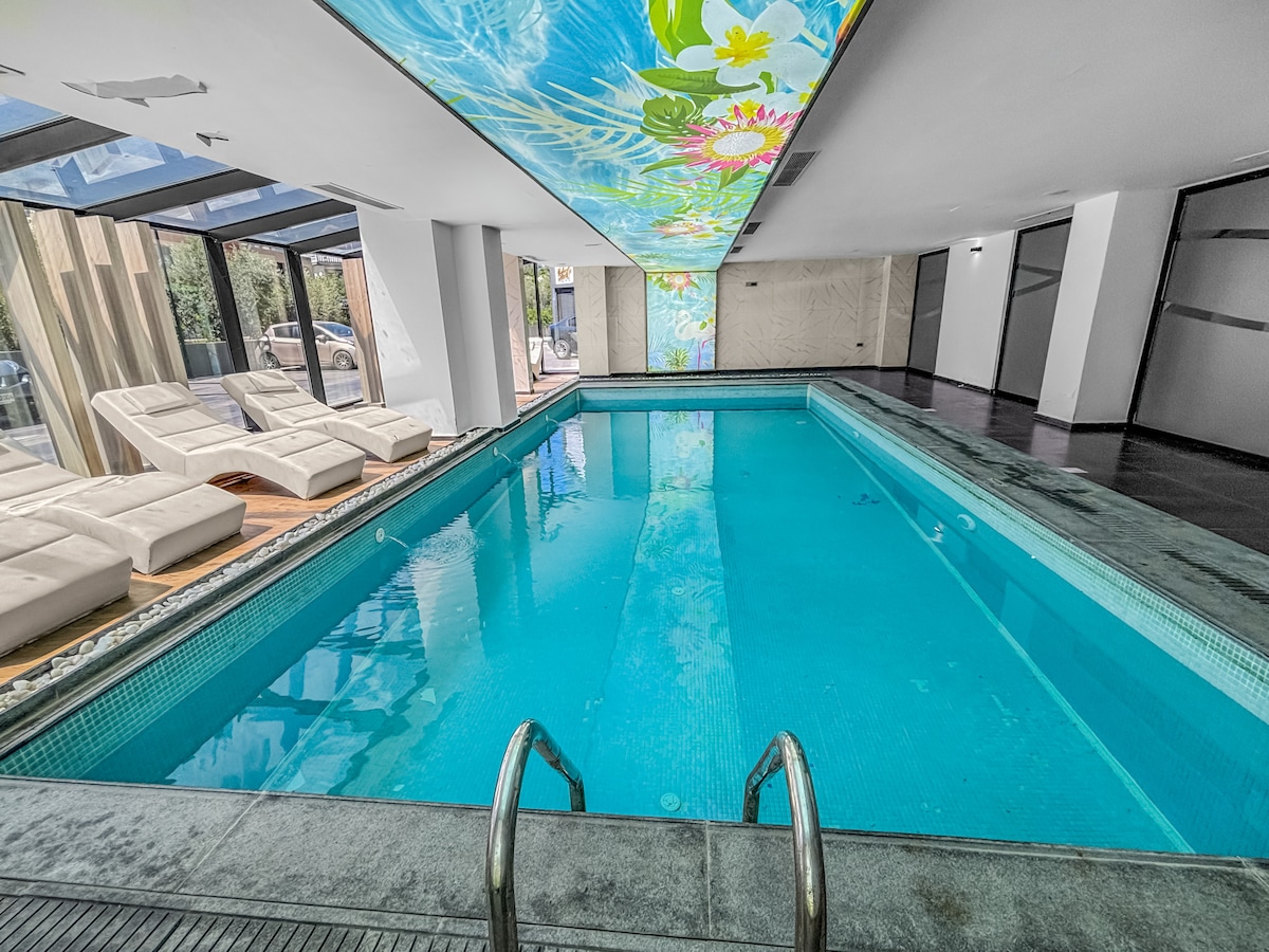 An indoor swimming pool features clear blue water, inviting relaxation. Sun loungers are positioned nearby, while a vibrant mural adorns the ceiling, adding color to the space. The surrounding area is well-lit, providing a cheerful atmosphere for leisure activities.