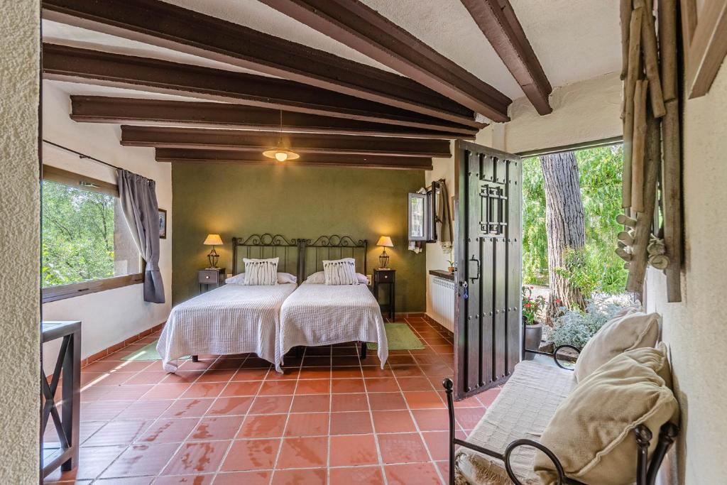 The room features two spacious twin beds with soft linens, accompanied by bedside lamps. Large windows offer views of greenery, while a cozy seating area is positioned near the entrance. Exposed wooden beams add character to the inviting space.