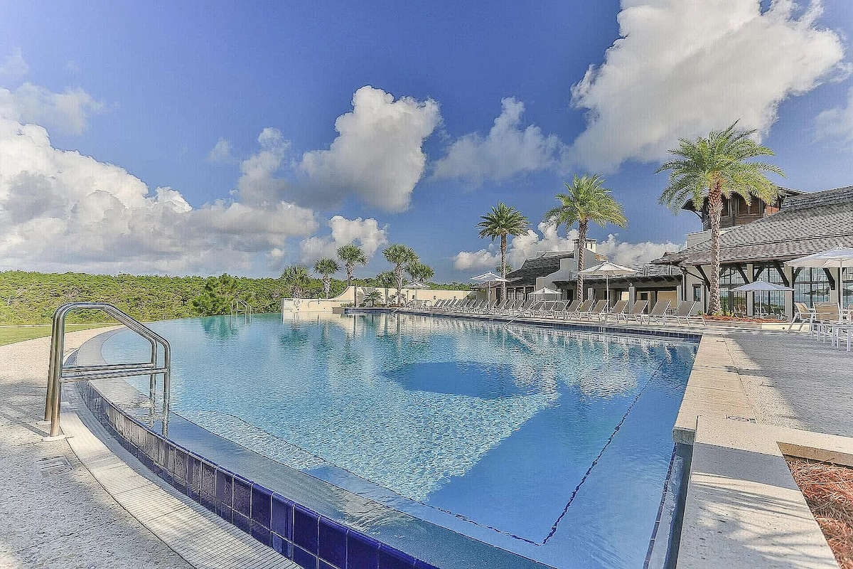 An expansive infinity pool reflects the blue sky and puffy clouds. Surrounding palm trees provide greenery, while lounge chairs line the pool deck. The adjacent clubhouse features shaded areas and a welcoming atmosphere, creating a serene setting for relaxation.