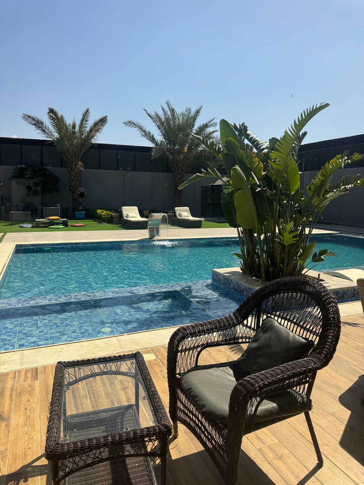 A private pool is framed by landscaped gardens and palm trees, reflecting a clear blue sky. Comfortable lounge chairs are positioned nearby on a wooden deck, accompanied by a wicker table. Water gently cascades from a fountain in the pool, enhancing the peaceful setting.