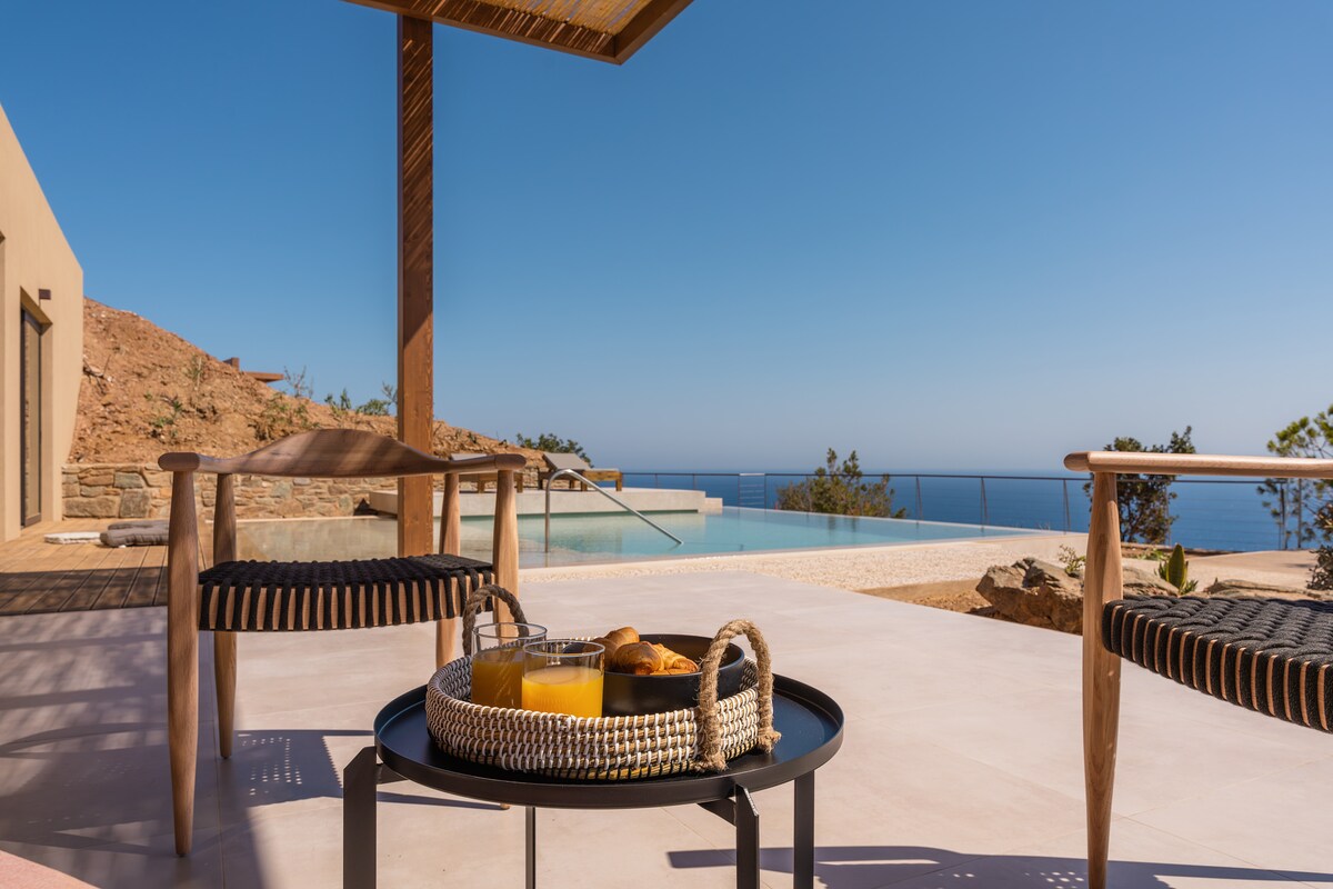 An outdoor seating area features two stylish chairs and a low table, holding a tray with drinks and pastries. In the background, a private swimming pool is visible, overlooking panoramic sea views under a clear blue sky.