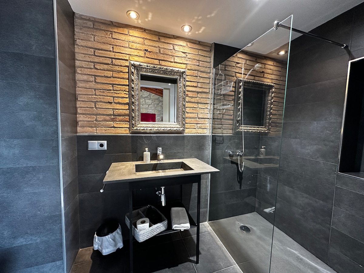 A modern bathroom features a walk-in shower with clear glass panels and a sleek black basin on a minimalist stand. Warm lighting highlights a stylish mirror above the basin, while textured brick walls add subtle character to the space.