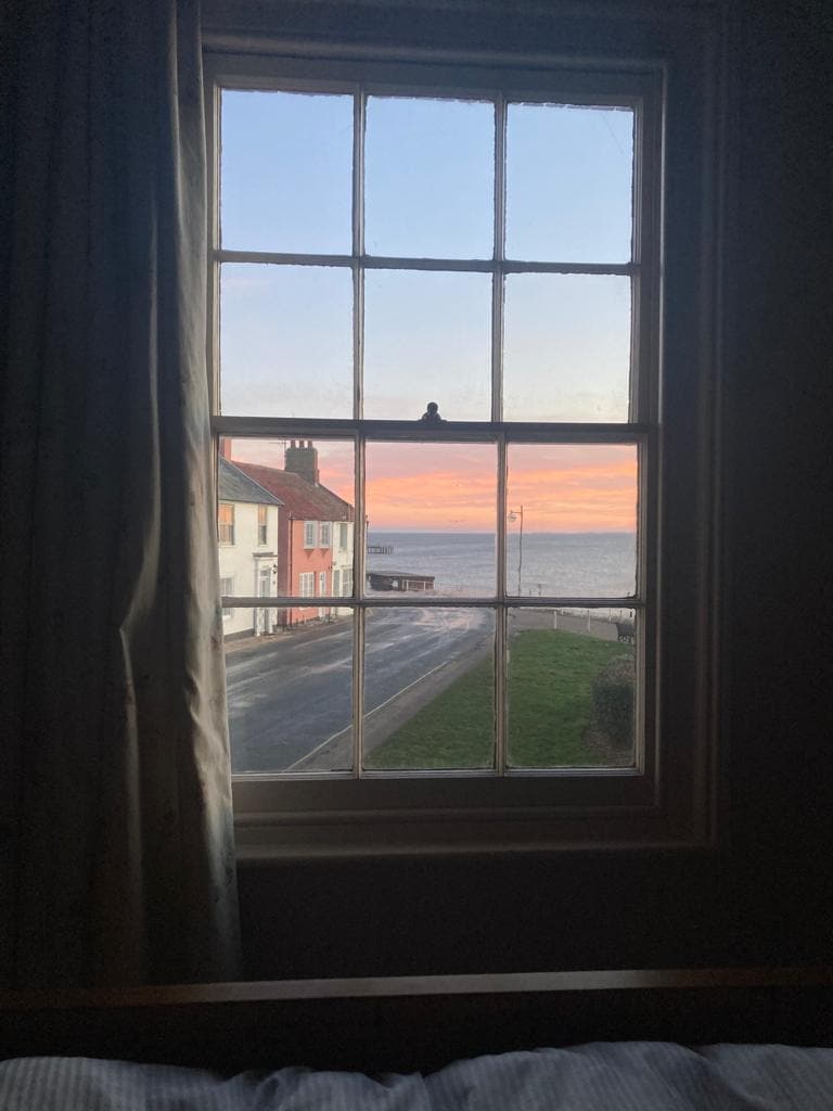 A large window frames a serene view of the sea at sunset, with soft pastel colors illuminating the horizon. Nearby houses along the street can be seen, enhancing the coastal ambiance. The curtains gently drape beside the window, allowing natural light to enter.