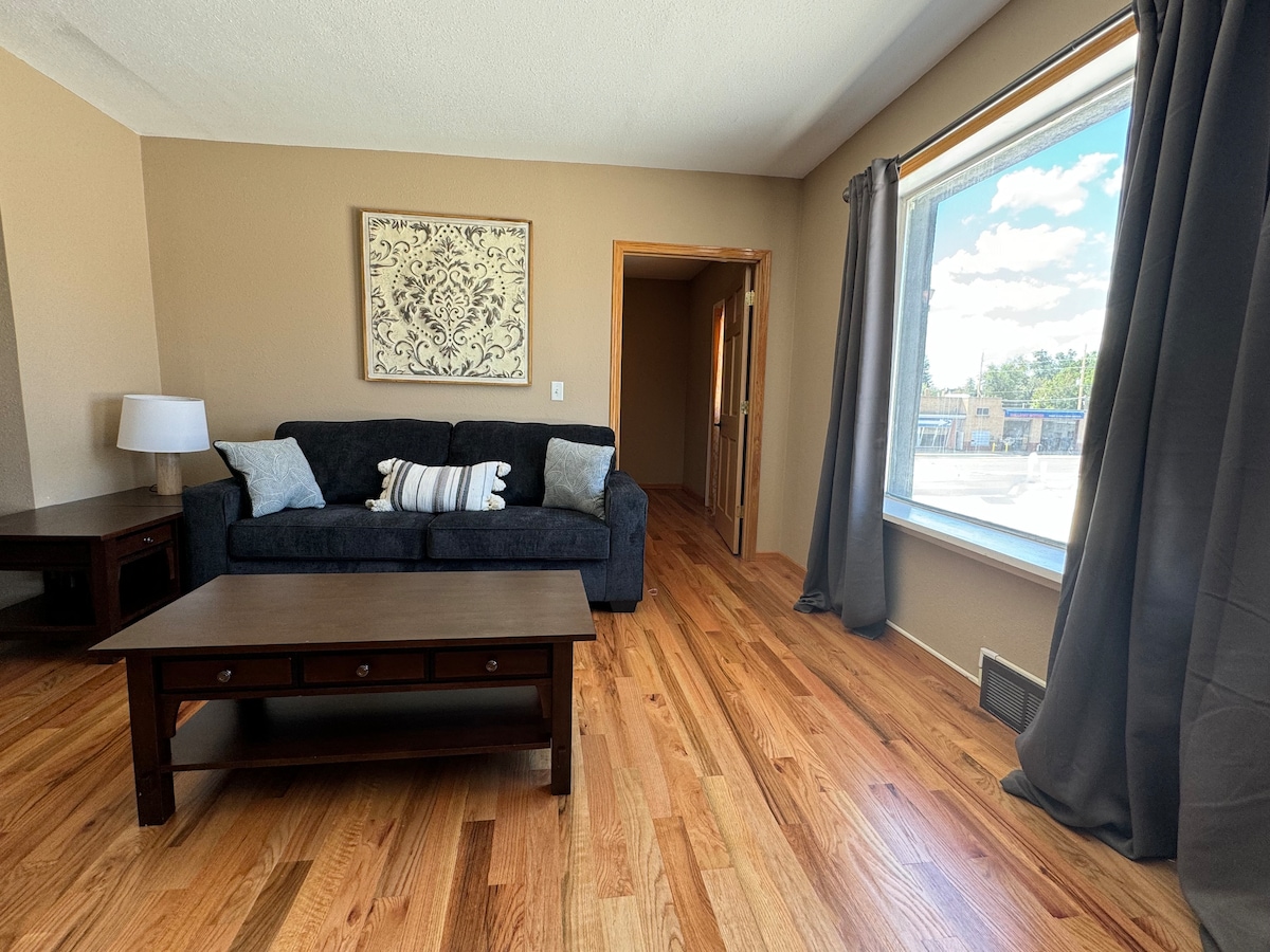 A living area is presented with hardwood flooring and a large window allowing natural light to fill the space. A dark sofa is paired with a wooden coffee table, and a decorative wall piece adds visual interest. Two curtains frame the window, while a doorway leads to another room.