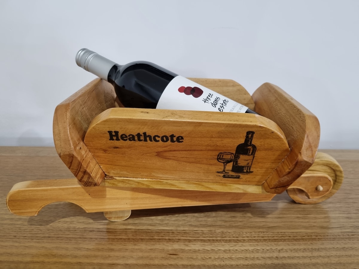 A wooden wine carrier, designed in a cart shape, showcases a bottle of red wine. The name 'Heathcote' is engraved on the side, adding a personal touch. The carrier rests atop a wooden surface, highlighting its craftsmanship and functionality for transporting beverages.