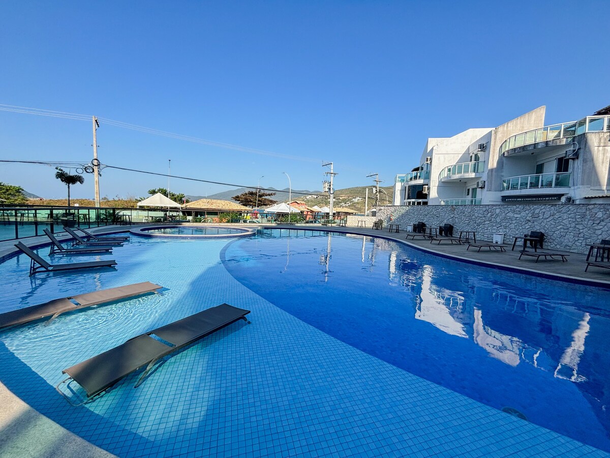 The pool area features a curved design with a crystal-clear water surface. Several lounge chairs are positioned along the poolside, offering relaxation spaces. Umbrellas provide shade in designated areas, and the surrounding landscape includes the modern architecture of the nearby condominium.